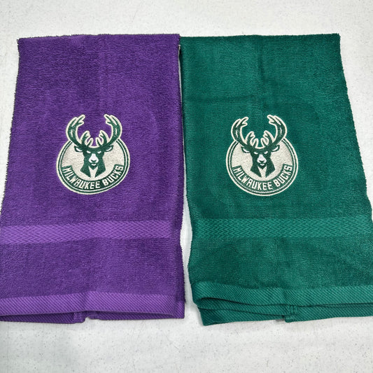Two folded towels side by side on a light gray surface, one purple and one green, both featuring a white Milwaukee Bucks logo and a horizontal textured stripe near the bottom.