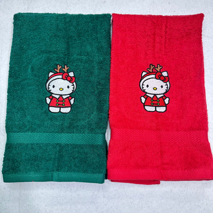 Two folded towels side by side on a light gray background. The left towel is dark green with a Hello Kitty holiday design, and the right towel is red with the same Hello Kitty holiday design.