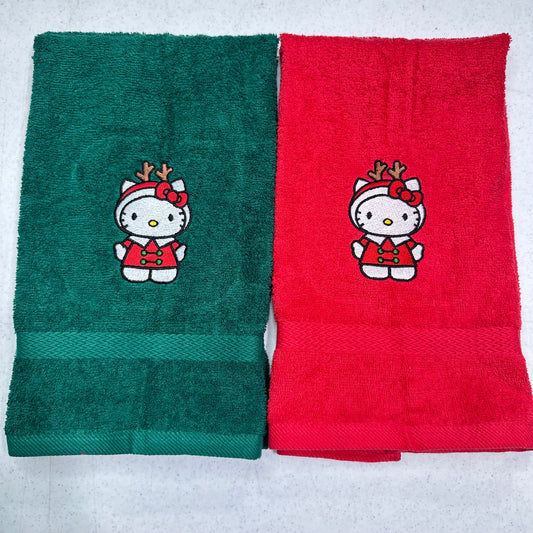 Two folded towels side by side on a light gray background. The left towel is dark green with a Hello Kitty holiday design, and the right towel is red with the same Hello Kitty holiday design.