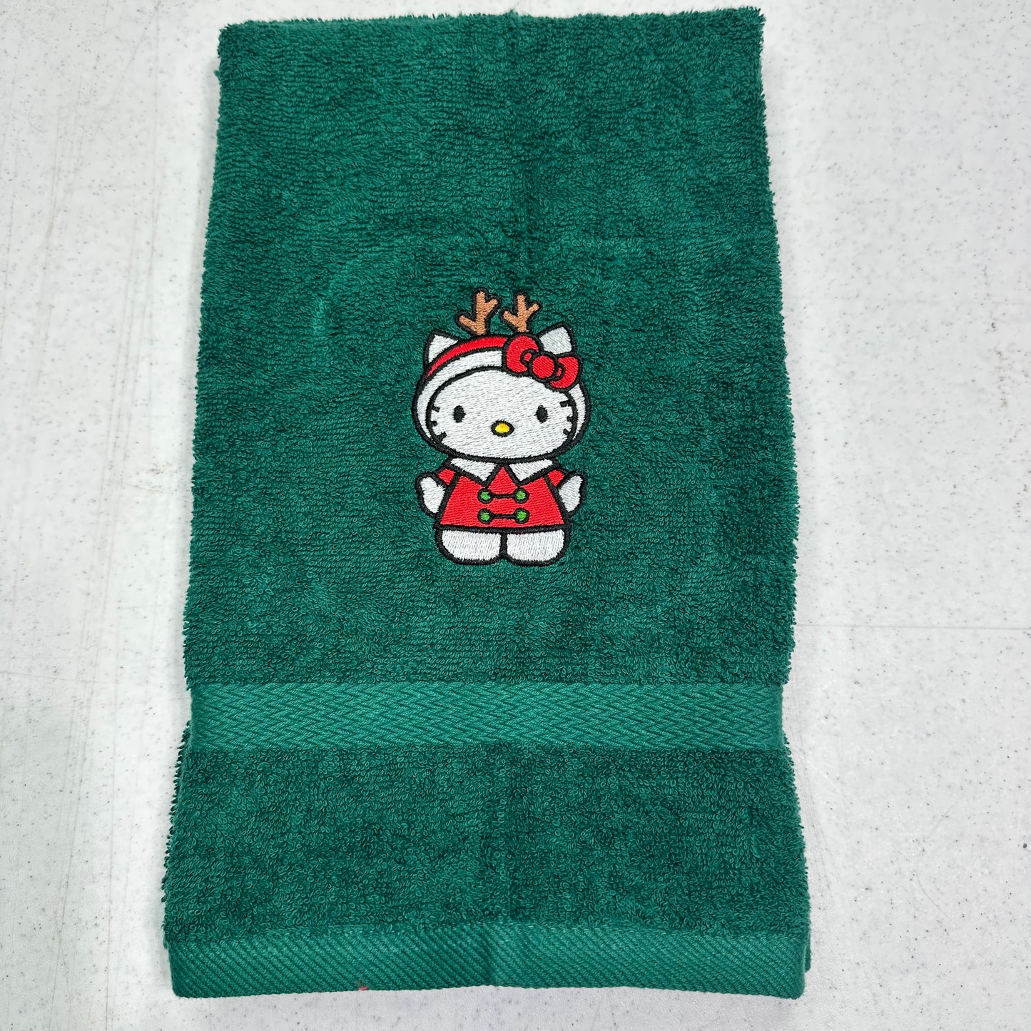 A dark green terry cloth towel featuring an embroidered Hello Kitty character in a red holiday outfit with reindeer antlers, laid flat on a light textured surface.