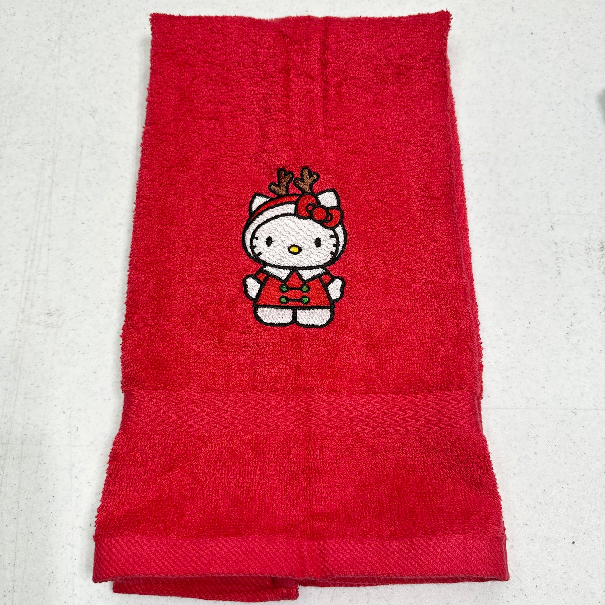 A folded red terry cloth towel featuring an embroidered Hello Kitty character wearing a holiday outfit with reindeer antlers, displayed on a light gray textured background.