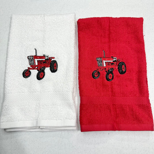 Two folded towels side by side on a light gray surface. The left towel is white with a red tractor embroidery, and the right towel is red with a matching red tractor embroidery.