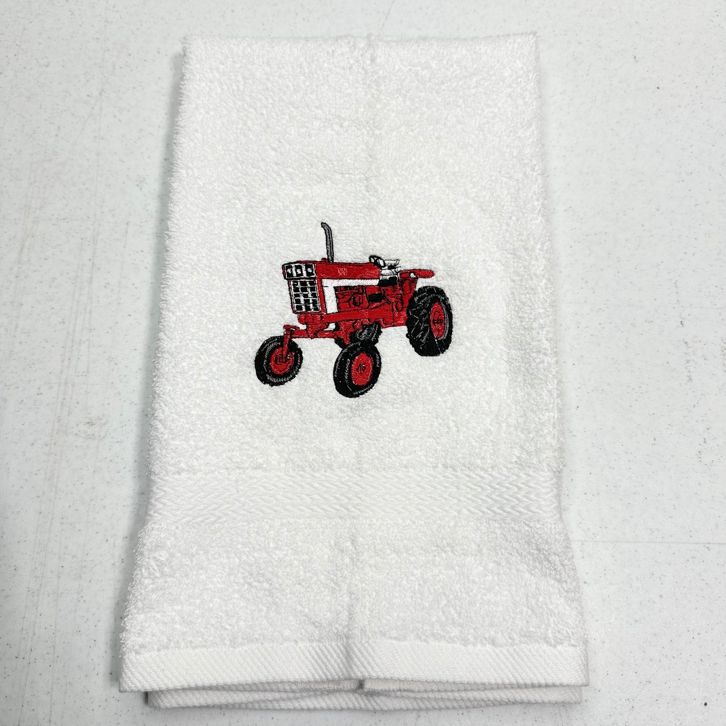 A folded white terry cloth towel featuring an embroidered red tractor design in the center, displayed on a light gray textured background.