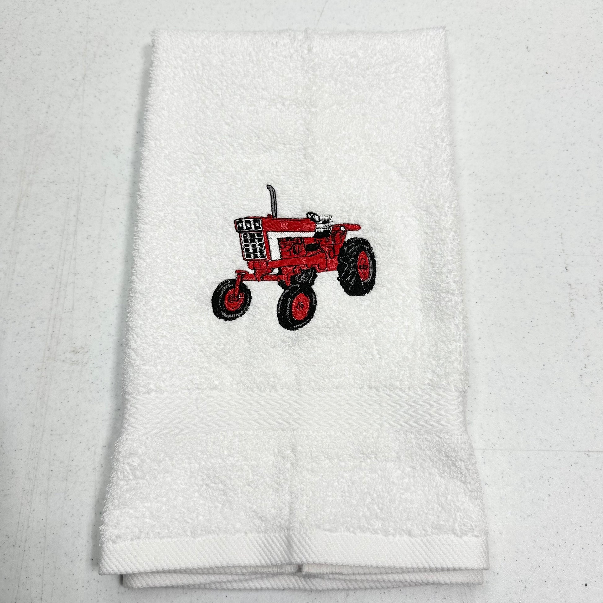 A folded white terry cloth towel featuring an embroidered red tractor design in the center, displayed on a light gray textured background.