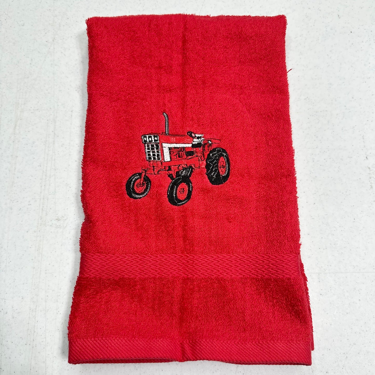 A red terry cloth hand towel featuring an embroidered red and black tractor design in the center, laid flat on a light gray textured background.