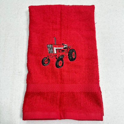 A red terry cloth hand towel featuring an embroidered red and black tractor design in the center, laid flat on a light gray textured background.