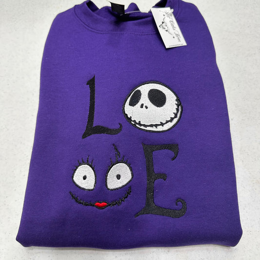 A folded purple sweatshirt featuring an embroidered design spelling 'LOVE' with Jack Skellington's face in for 'O' and a smiling face with large eyes and red lips for 'V'. A white hangtag is attached at the collar.