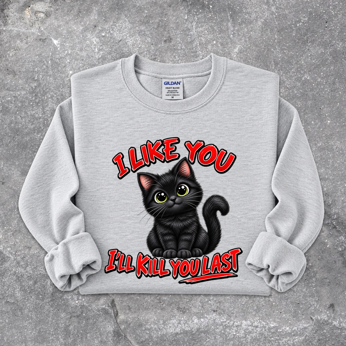 A light gray crewneck sweatshirt featuring a graphic of a black cat with green eyes and the text 'I LIKE YOU I'LL KILL YOU LAST' in red and black, laid flat on a textured gray concrete background. The Gildan brand tag is visible inside the collar.
