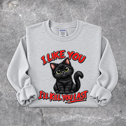 A light gray crewneck sweatshirt featuring a graphic of a black cat with green eyes and the text 'I LIKE YOU I'LL KILL YOU LAST' in red and black, laid flat on a textured gray concrete background. The Gildan brand tag is visible inside the collar.
