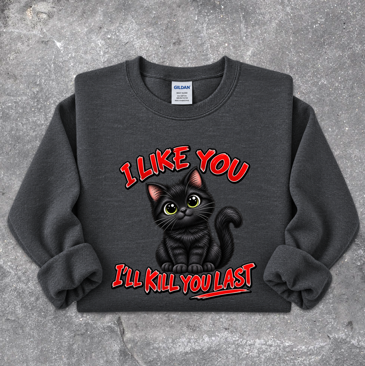 A dark gray crewneck sweatshirt featuring a graphic of a black cat with yellow eyes and the text 'I LIKE YOU I'LL KILL YOU LAST' in red, laid flat on a textured gray concrete background. The neck label shows the brand Gildan.