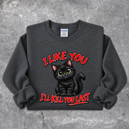A dark gray crewneck sweatshirt featuring a graphic of a black cat with yellow eyes and the text 'I LIKE YOU I'LL KILL YOU LAST' in red, laid flat on a textured gray concrete background. The neck label shows the brand Gildan.