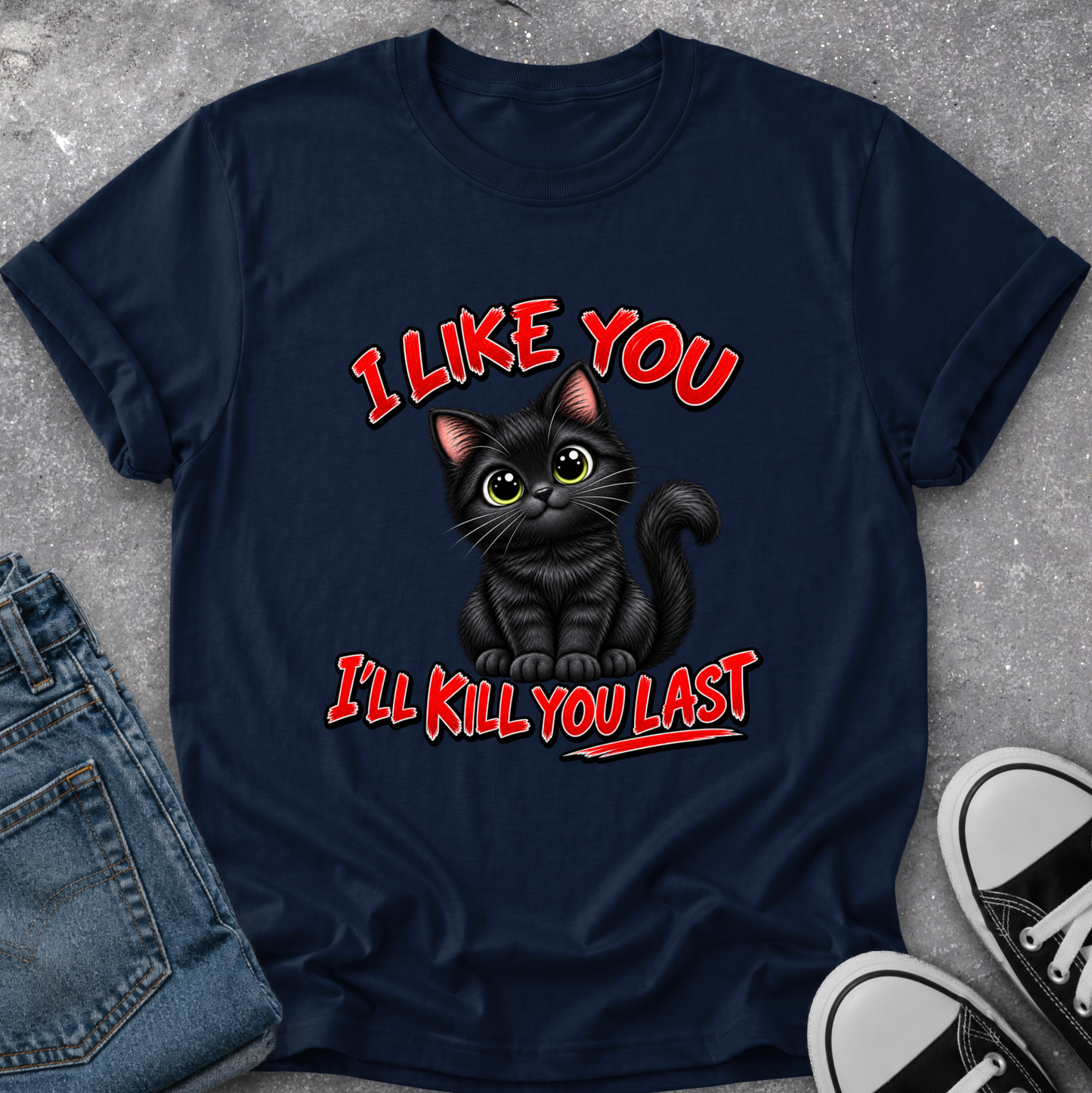A navy blue graphic t-shirt laid flat on a gray concrete surface, styled with folded blue jeans on the left and black-and-white sneakers on the right. The t-shirt features a cartoon illustration of a black cat with green eyes and the text 'I LIKE YOU I'LL KILL YOU LAST' in red and white.