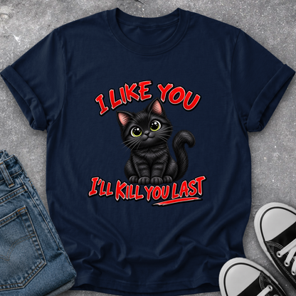 A navy blue graphic t-shirt laid flat on a gray concrete surface, styled with folded blue jeans on the left and black-and-white sneakers on the right. The t-shirt features a cartoon illustration of a black cat with green eyes and the text 'I LIKE YOU I'LL KILL YOU LAST' in red and white.