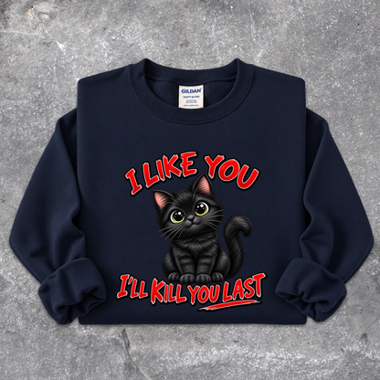 A navy blue crewneck sweatshirt featuring a graphic of a black cat with green eyes and the text 'I LIKE YOU I'LL KILL YOU LAST' in red with a black outline, laid flat on a textured gray concrete background. The Gildan brand label is visible inside the collar.