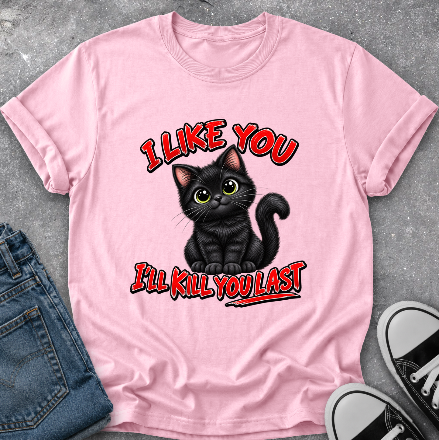 A light pink short-sleeve graphic T-shirt laid flat on a gray textured surface, styled with folded blue jeans and black-and-white sneakers. The shirt features a black cartoon cat with green eyes and red outlined text reading 'I LIKE YOU I'LL KILL YOU LAST'.
