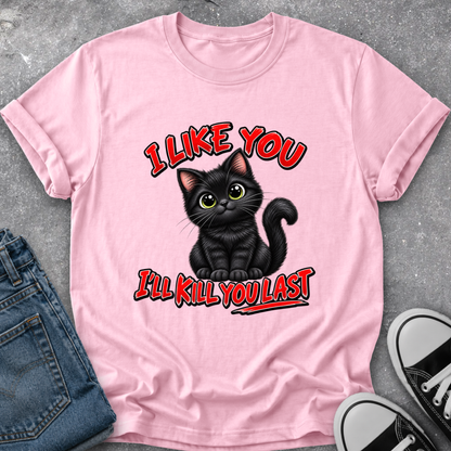 A light pink short-sleeve graphic T-shirt laid flat on a gray textured surface, styled with folded blue jeans and black-and-white sneakers. The shirt features a black cartoon cat with green eyes and red outlined text reading 'I LIKE YOU I'LL KILL YOU LAST'.