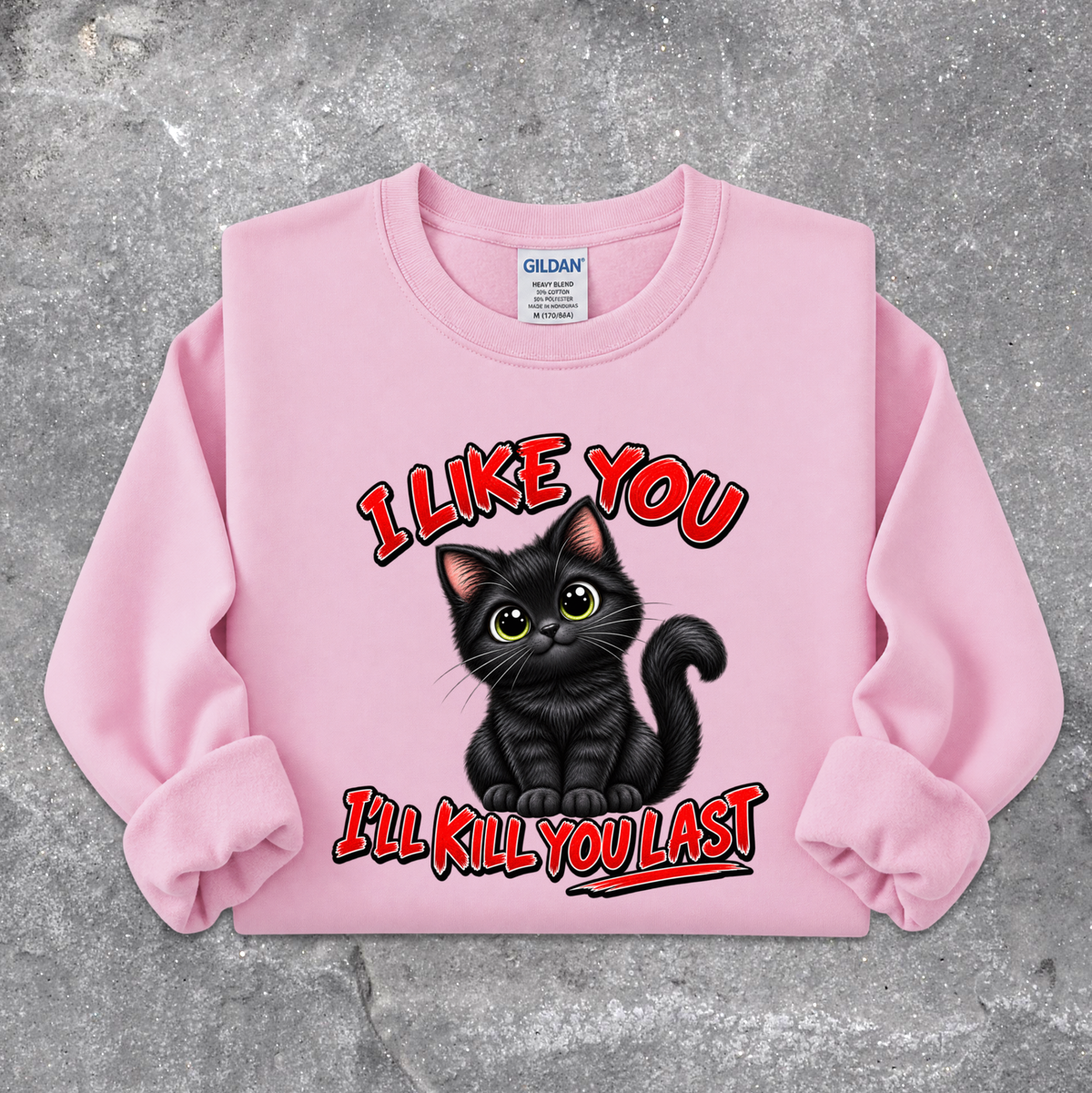 A light pink crewneck sweatshirt featuring a graphic of a black cat with green eyes and the text 'I LIKE YOU I'LL KILL YOU LAST' in red with black outlines, laid flat on a textured gray concrete surface. The Gildan brand tag is visible inside the collar.