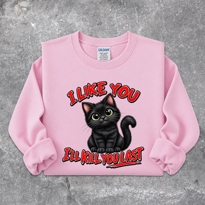 A light pink crewneck sweatshirt featuring a graphic of a black cat with green eyes and the text 'I LIKE YOU I'LL KILL YOU LAST' in red with black outlines, laid flat on a textured gray concrete surface. The Gildan brand tag is visible inside the collar.