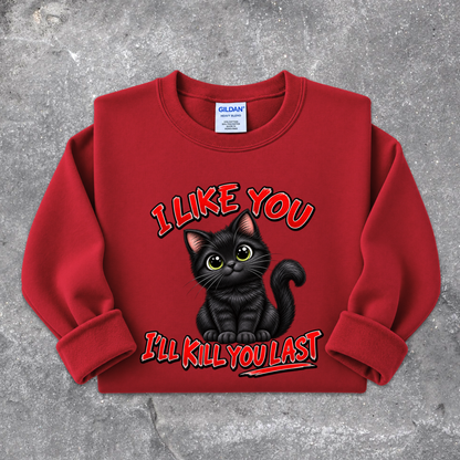 A red crewneck sweatshirt featuring a graphic of a black cat with the text 'I LIKE YOU I'LL KILL YOU LAST' in red and black, laid flat on a textured gray background. The neck label shows Gildan.
