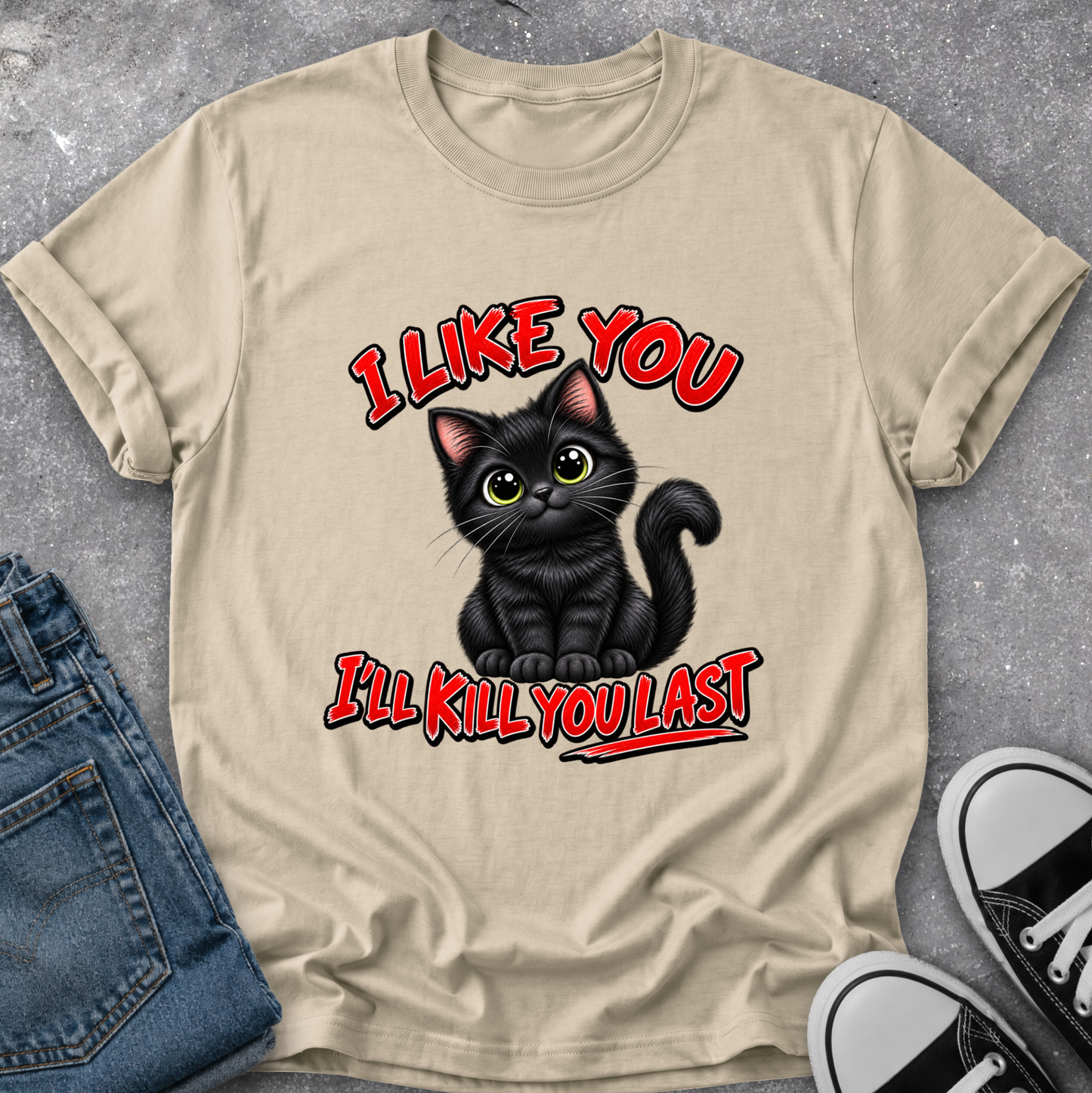 A beige graphic t-shirt laid flat on a gray concrete surface, styled with a pair of blue jeans and black-and-white sneakers. The t-shirt features a cartoon illustration of a black cat with green eyes and the text 'I LIKE YOU I'LL KILL YOU LAST' in red with black and white outlines.