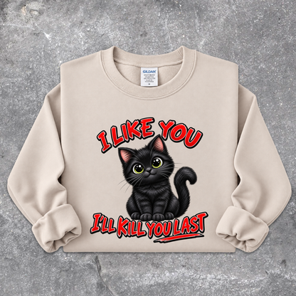 A beige crewneck sweatshirt featuring a graphic of a black cat with green eyes and the text 'I LIKE YOU I'LL KILL YOU LAST' in red and black, laid flat on a gray concrete-textured background. The Gildan brand tag is visible inside the collar.