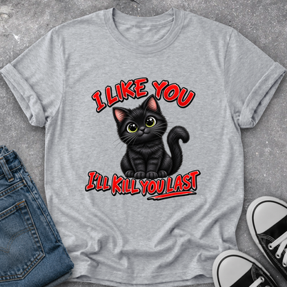 A heather gray graphic t-shirt featuring a cartoon black cat with green eyes and the text 'I LIKE YOU I'LL KILL YOU LAST' in red with black outlines. The t-shirt is styled in a flat-lay arrangement with blue jeans and black-and-white sneakers on a gray concrete-textured background.