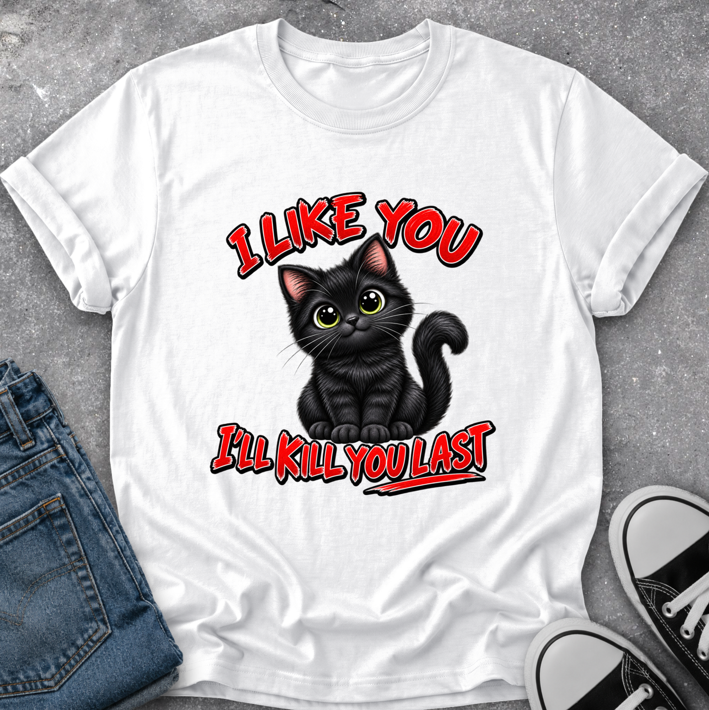 A white short-sleeve graphic T-shirt laid flat on a gray textured surface, styled with blue jeans and black-and-white sneakers. The shirt features a cartoon black cat with green eyes and red text reading 'I LIKE YOU I'LL KILL YOU LAST'.
