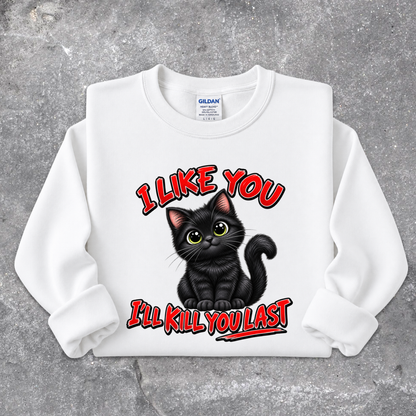 A white long-sleeve crewneck sweatshirt with a graphic of a black cat and the text 'I LIKE YOU I'LL KILL YOU LAST' in red and black, displayed flat on a gray concrete-textured background. The neck label shows Gildan.