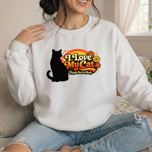 A woman wearing a white crewneck sweatshirt with a graphic that reads 'I Love My Cat People Not So Much' alongside a black cat silhouette and orange flowers, paired with light blue ripped jeans in a cozy indoor setting.