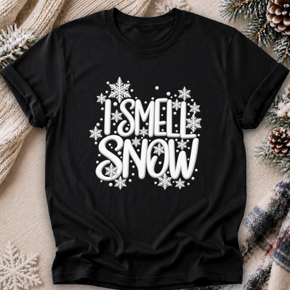 A black short-sleeve crewneck t-shirt with a white graphic reading 'I SMELL SNOW' surrounded by snowflakes, laid flat on a cozy holiday-themed background with a knit sweater, pinecones, and plaid fabric.