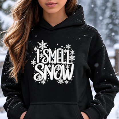 A woman wearing a black pullover hoodie with a white graphic that reads 'I SMELL SNOW' surrounded by snowflakes, photographed outdoors in the snow.