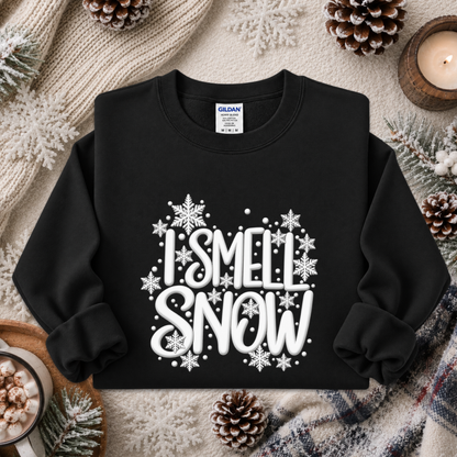 A black crewneck sweatshirt with a white graphic reading 'I SMELL SNOW' surrounded by snowflakes, styled in a cozy flat lay with pinecones, a knit blanket, and a candle.