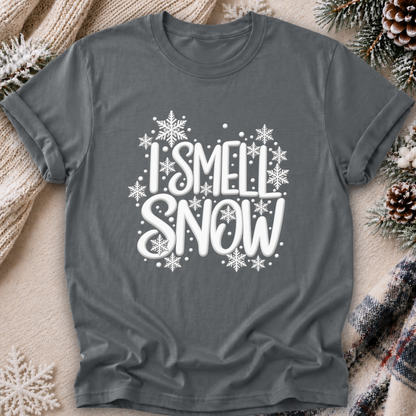 A dark gray short-sleeve t-shirt with a white graphic reading 'I SMELL SNOW' surrounded by snowflakes, laid flat on a cozy holiday-themed background with knitted blankets, pinecones, and evergreen branches.