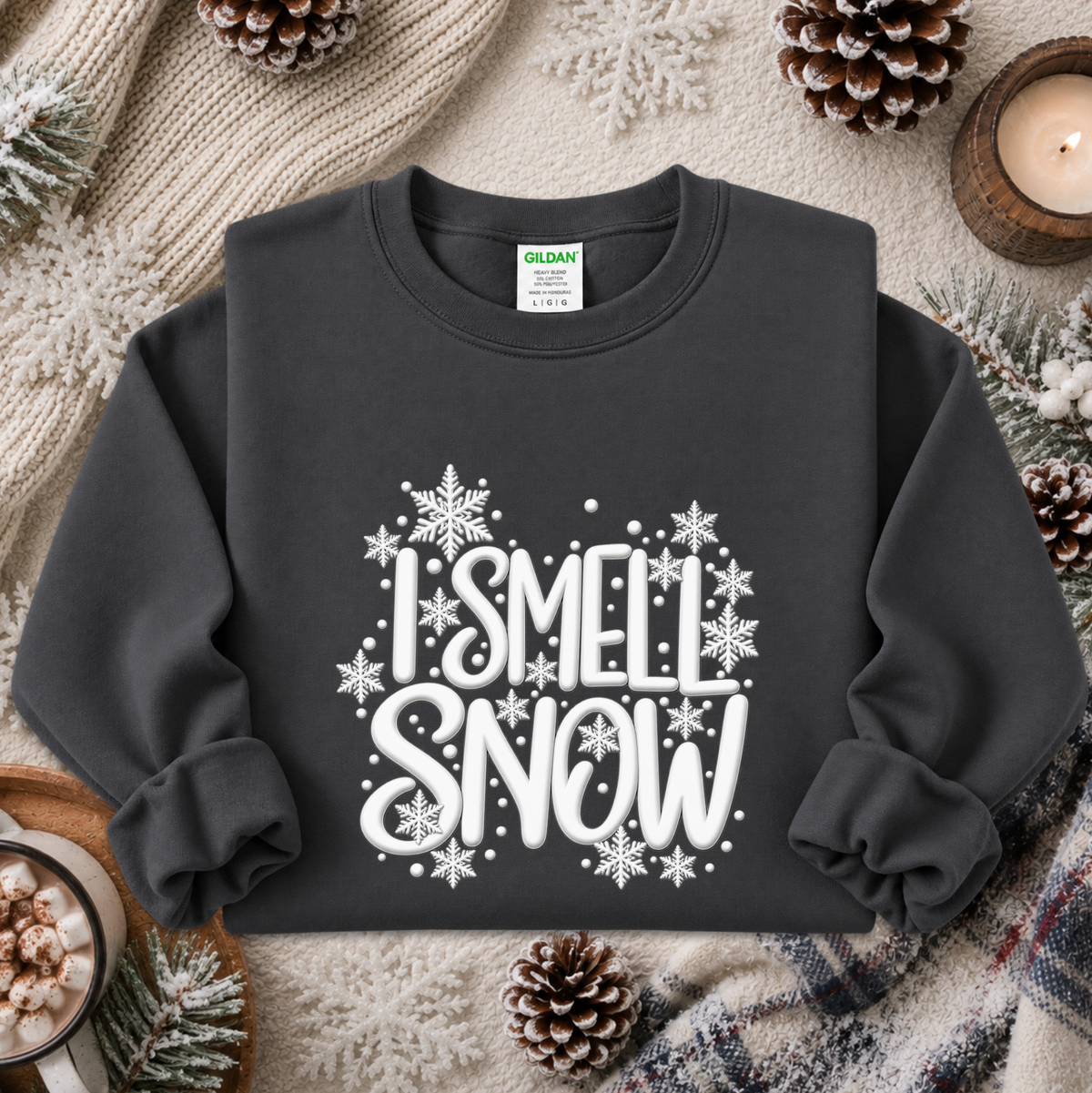 A dark charcoal gray sweatshirt with a white graphic reading 'I SMELL SNOW' surrounded by snowflakes, styled in a cozy flat lay with pinecones, a candle, and a knit blanket.