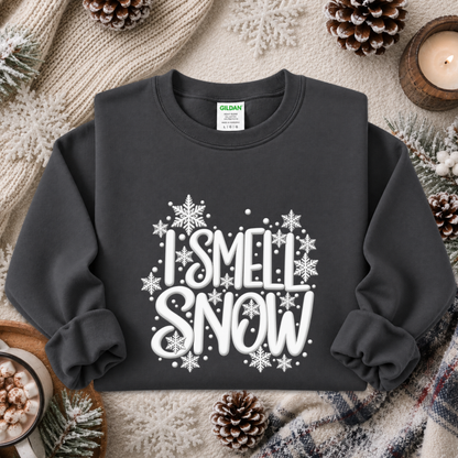 A dark charcoal gray sweatshirt with a white graphic reading 'I SMELL SNOW' surrounded by snowflakes, styled in a cozy flat lay with pinecones, a candle, and a knit blanket.