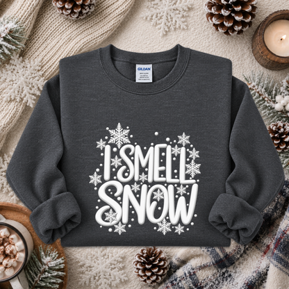 A dark charcoal gray sweatshirt with a white graphic reading 'I SMELL SNOW' surrounded by snowflakes, styled in a cozy flat lay with pinecones, a candle, and knitted blankets.