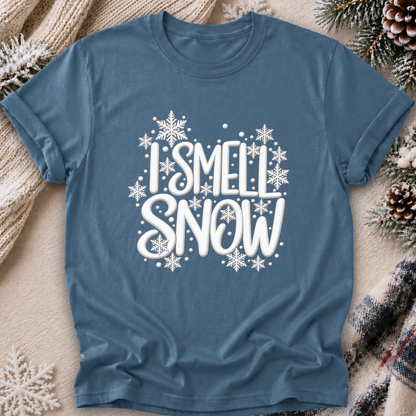 A slate blue short-sleeve t-shirt with a white graphic reading 'I SMELL SNOW' surrounded by white snowflakes, laid flat on a cozy beige blanket with pinecones and knit textures around it.
