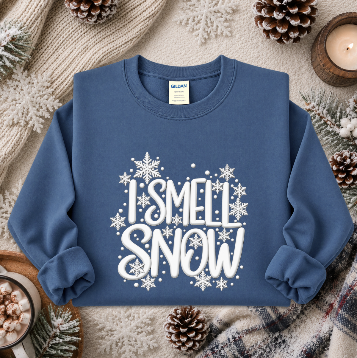 A blue crewneck sweatshirt with a white graphic reading 'I SMELL SNOW' surrounded by snowflakes, styled in a cozy flat lay with pinecones, a candle, and knitted blankets.