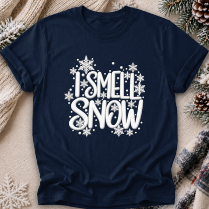 A navy blue short-sleeve t-shirt with a white graphic reading 'I SMELL SNOW' surrounded by snowflakes, laid flat on a cozy beige blanket with pine branches and a plaid scarf.