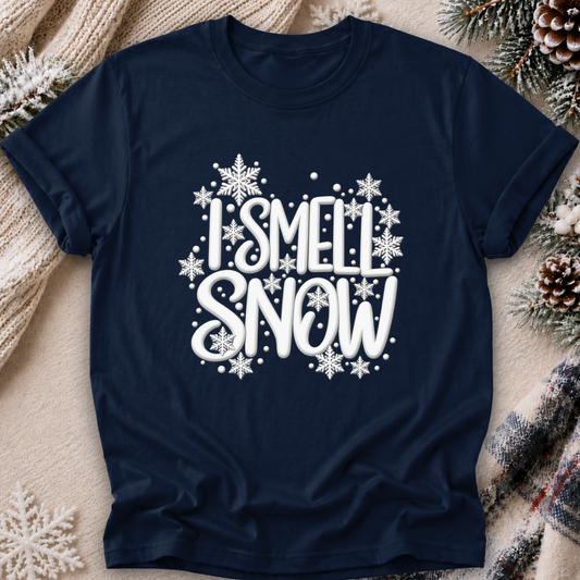 A navy blue short-sleeve t-shirt with a white graphic reading 'I SMELL SNOW' surrounded by snowflakes, laid flat on a cozy beige blanket with pine branches and a plaid scarf.