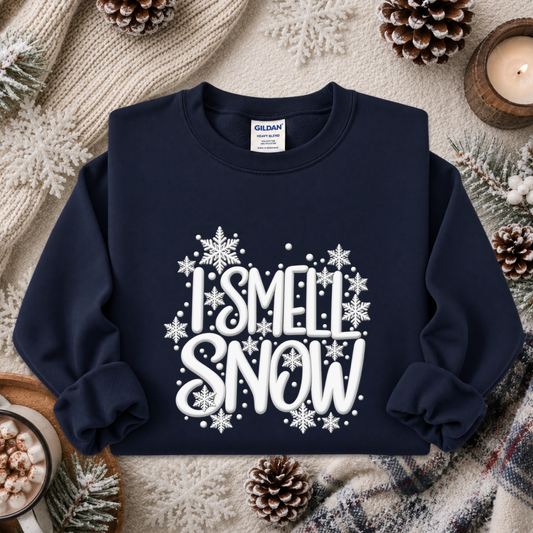 A navy blue crewneck sweatshirt with a white graphic reading 'I SMELL SNOW' surrounded by snowflakes, styled in a cozy flat lay with a knit blanket, pinecones, and a candle.