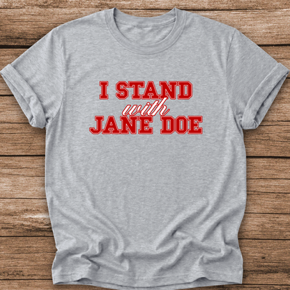 A light heather gray t-shirt with the slogan 'I STAND with JANE DOE' printed in red across the chest, laid flat on a rustic brown wooden plank background.