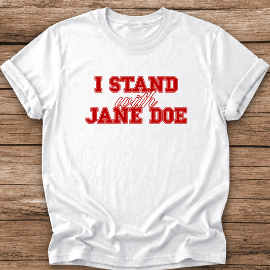 A white short-sleeve crewneck t-shirt with red text reading 'I STAND with JANE DOE' printed on the front, laid flat on a rustic brown wooden plank background.