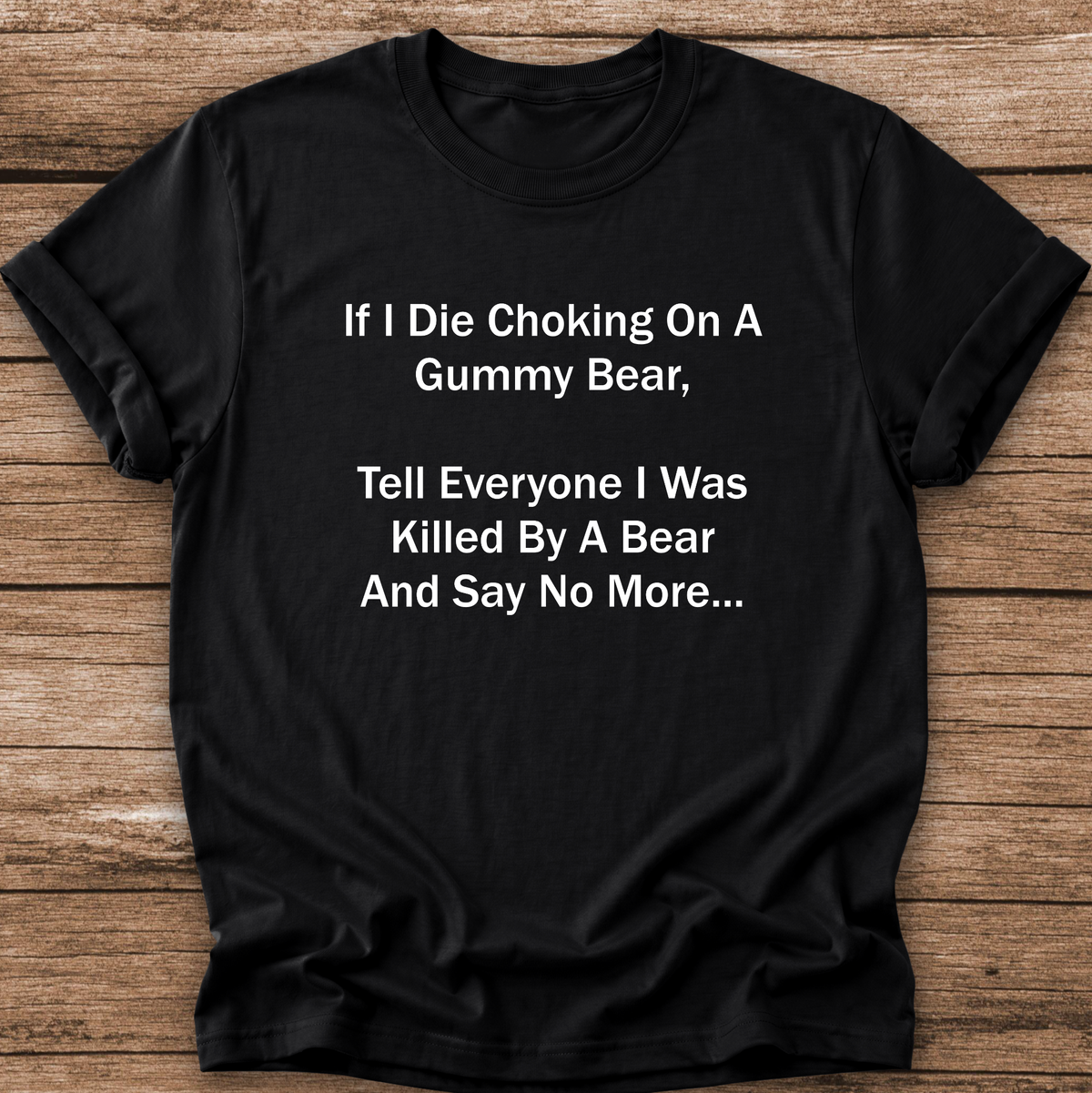 A black short-sleeve t-shirt with white printed text on the front, laid flat on a rustic wooden background. The text reads: 'If I Die Choking On A Gummy Bear, Tell Everyone I Was Killed By A Bear And Say No More.