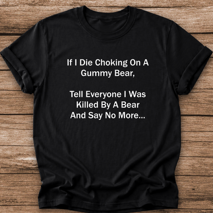A black short-sleeve t-shirt with white printed text on the front, laid flat on a rustic wooden background. The text reads: 'If I Die Choking On A Gummy Bear, Tell Everyone I Was Killed By A Bear And Say No More.