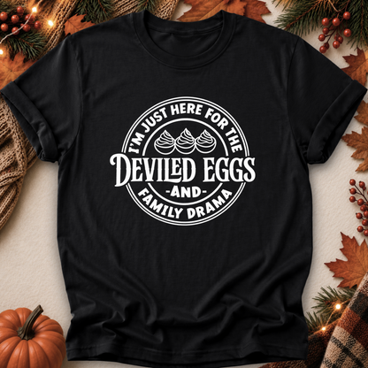 A black short-sleeve graphic T-shirt laid flat on a neutral surface with autumn-themed props including a pumpkin, pine branches, leaves, and string lights. The shirt has a large white circular print reading “I’m Just Here For the Deviled Eggs and Family Drama” with three deviled eggs illustrated in the center.