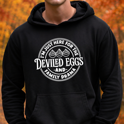 A person wearing a black pullover hoodie with a white circular graphic that reads “I’m Just Here For the Deviled Eggs and Family Drama” with two deviled eggs illustration, set against a blurred autumn foliage background.