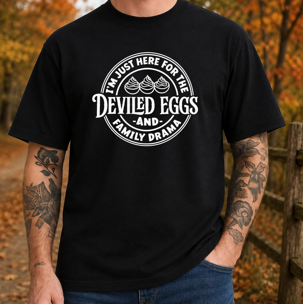 A man wearing a black graphic t-shirt with a white circular print that reads 'I'M JUST HERE FOR THE DEVILED EGGS AND- FAMILY DRAMA' with three deviled eggs illustrations, set against an autumn outdoor background.