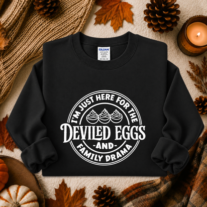 A black crewneck sweatshirt with a white graphic print reading 'I'M JUST HERE FOR THE DEVILED EGGS AND FAMILY DRAMA' laid flat on a cozy autumn-themed background with pinecones, leaves, a candle, and a knit blanket.