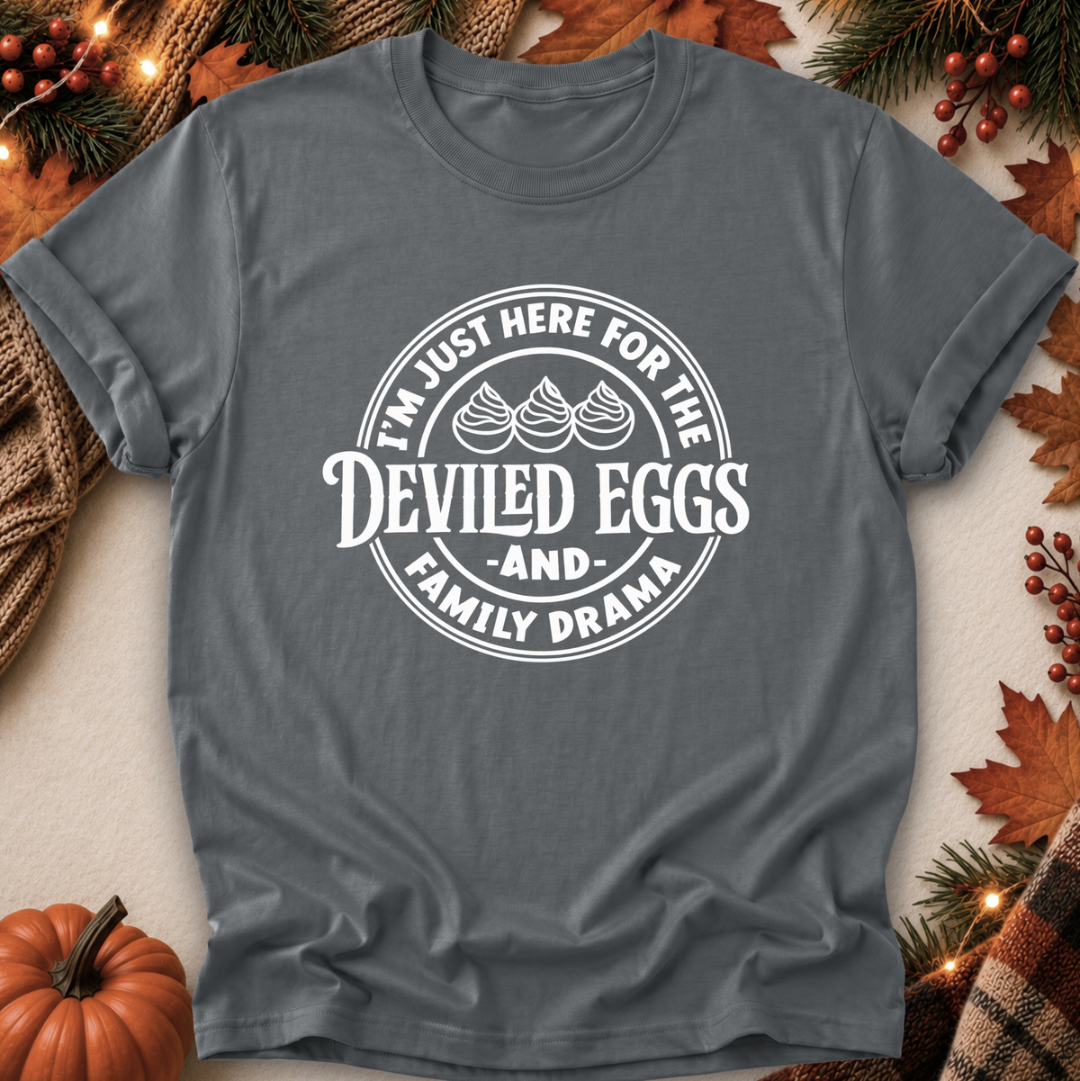 A dark gray crewneck t-shirt with a white circular graphic that reads 'I'M JUST HERE FOR THE DEVILED EGGS AND FAMILY DRAMA' with three egg illustrations, displayed in a flat-lay autumn setting with pumpkins, pine branches, and leaves.